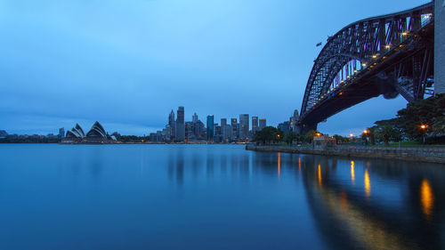 Illuminated bridge over river by buildings against sky at dusk