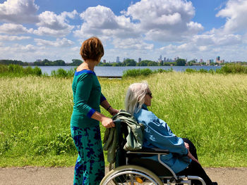 Young woman and elderly lady in wheelchair walking along a lake, looking at skyline in the distance