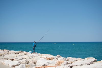 Rear view of man fishing by sea against clear blue sky