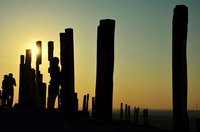 Silhouette people standing on wooden posts in sea against clear sky during sunset