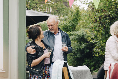 Smiling senior friends standing with arm around enjoying garden party at back yard