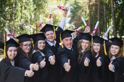 A group of graduates in robes give a thumbs up outdoors. elderly student