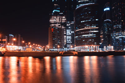 Illuminated buildings by river against sky at night