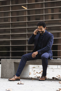 Businessman talking on smart phone while sitting on concrete in city