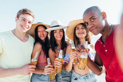 Friends holding drinks while standing at beach against sky