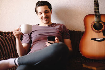 Happy young man using smart phone while drinking tea at home