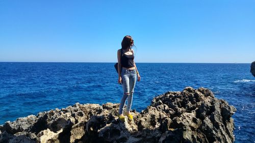 Young woman standing on rock by sea against clear sky / woman on rocky beach