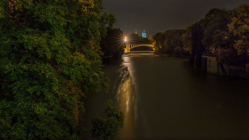 Illuminated river amidst trees against sky at night