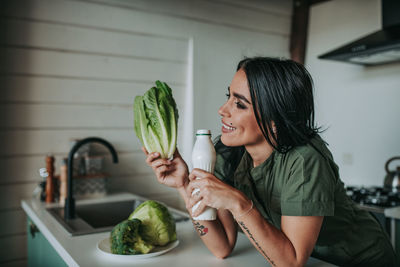 Smiling woman drinking milk and eating vegetables in kitchen at home