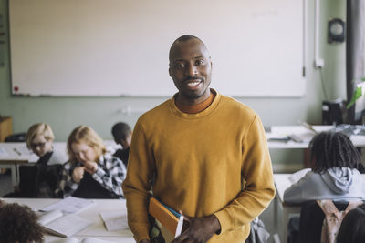 Portrait of male teacher with books standing in classroom