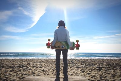 Full length rear view of woman holding skateboard while standing on beach