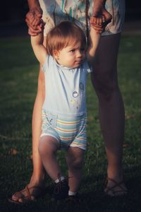 Baby boy with blonde hair and blue eyes on grass making first steps helped by parent arms