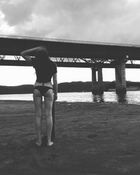 Rear view of seductive woman standing at beach against bridge