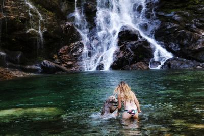 Rear view of woman playing with dog in lake against waterfall