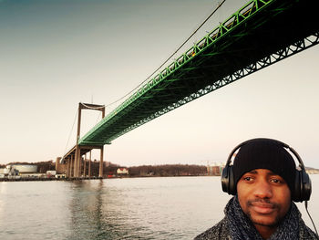 Portrait of man listening music under bridge in city against sky