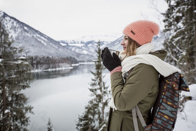 Young woman with hot drink standing in alpine winter landscape with lake