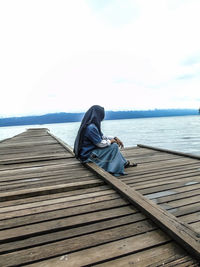 Side view of man sitting on pier over sea against sky