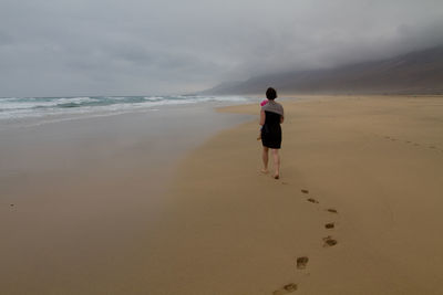 Rear view of woman leaving footprints at beach against cloudy sky
