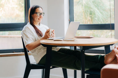 Young laughing female freelancer in glasses drinking coffee and working on laptop at home.