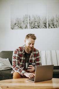 Man with beard holds cup of coffee while working on laptop computer