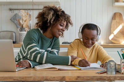 Happy smiled black child boy with pleased mother sit at desk with pencil write at school tutorial