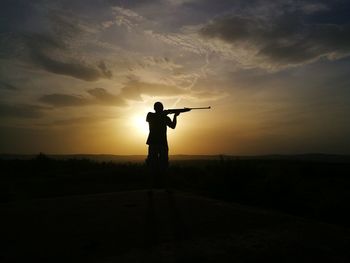 Silhouette man shooting rifle on field against sky during sunset