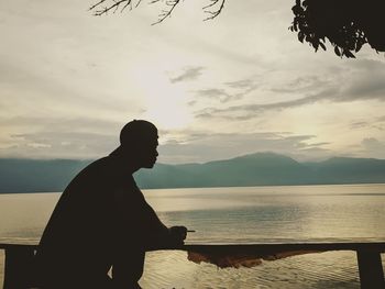 Side view of silhouette man looking at lake against sky during sunset