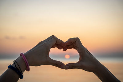 Close-up of hand over heart shape against sky during sunset