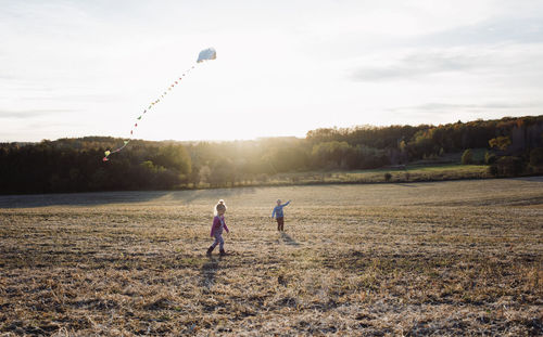 Brother flying kite while sister walking on field against sky