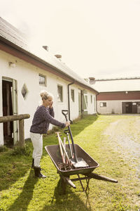 Female farmer using phone while holding equipment over wheelbarrow at farm