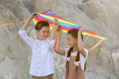 A girl and a boy carry a bright colored kite over their heads while walking in the mountains.