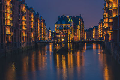 Illuminated buildings by river against sky at dusk