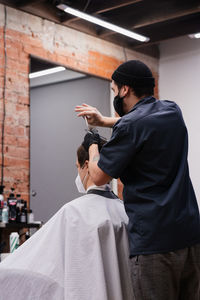 Barber in protective mask and latex glove styling adult man in a barbershop salon. new normal