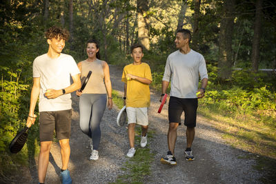 Happy family with tennis ball and rackets walking on dirt road in forest during vacation