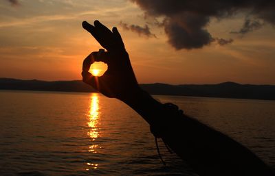 Silhouette person hand holding sea against sky during sunset