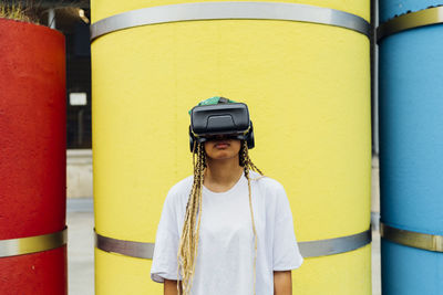 Young woman wearing virtual reality simulator standing in front of yellow concrete pipe