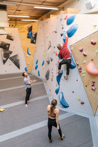 High angle view of male and female students looking at mature coach climbing wall in gym