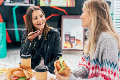 Two women are chatting and eating fast food at the street market, woman using audio messenger.