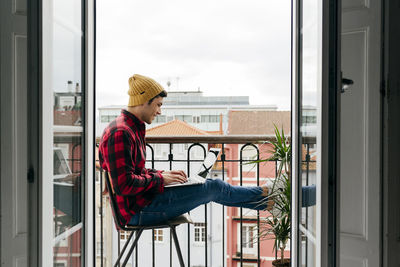 Side view of trendy focused caucasian man with beanie hat and shirt sitting on chair at balcony while working remotely on laptop at home