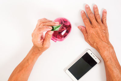 Close-up of man inserting money in piggy bank with mobile phone against white background