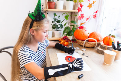 Halloween. a teenage girl in a witch hat drawing a pumpkin sitting by the window