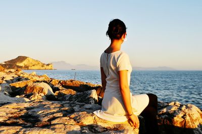 Rear view of woman sitting on rock at beach during sunset