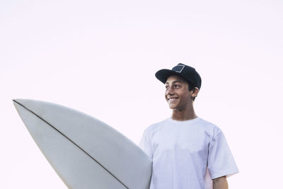 Boy looking away while holding surfboard against clear sky