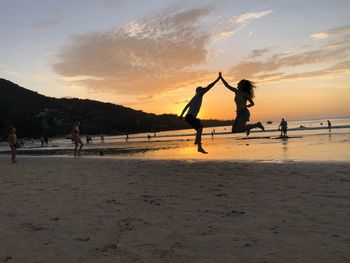 Silhouette couple giving high-five while jumping at beach against sky during sunset