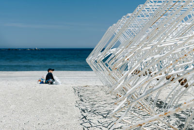 Man sitting at beach by metallic structure against sky