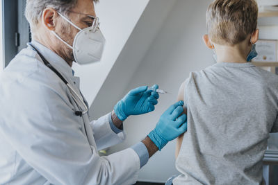 Doctor wearing protective face mask giving covid-19 vaccine to boy while standing at clinic