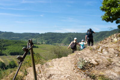 Metal railing on the vineyard trail, dry slate rocks visible and family in the background.