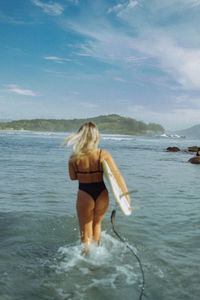 Rear view of woman in bikini standing in sea against sky