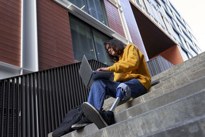 Young man with artificial limb using laptop while sitting on steps against building