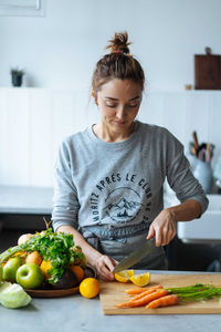 Adult lady slicing fresh lemon near ripe fruits and vegetables while standing in kitchen and preparing healthy dish for lunch